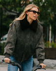 A woman riding a bicycle outdoors wearing a dark charcoal gray suede jacket with a black faux-fur collar and blue jeans.