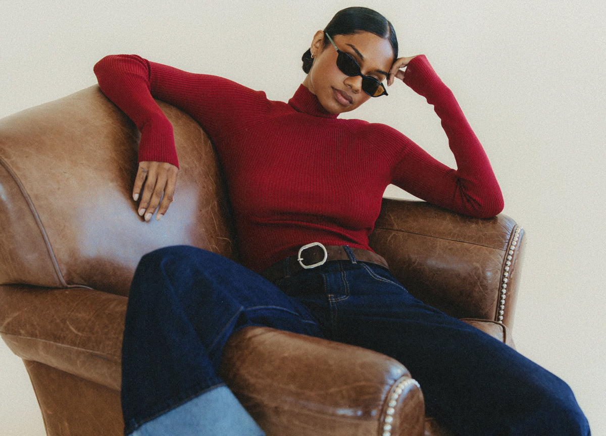 A woman wearing a fitted red ribbed turtleneck sweater and dark blue jeans sits in a brown leather armchair against a plain light background.