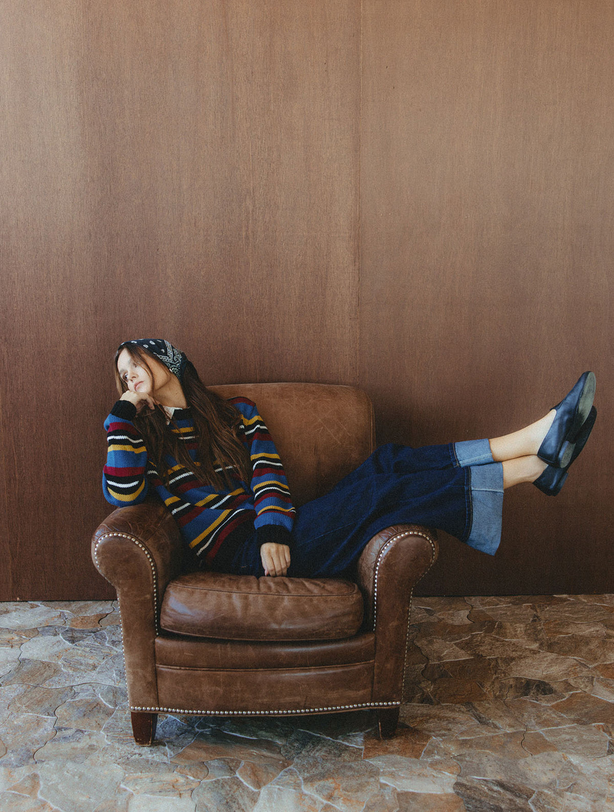A woman reclining in a brown leather armchair against a wood-paneled wall, wearing a colorful striped knit sweater, dark blue pants, and blue shoes.